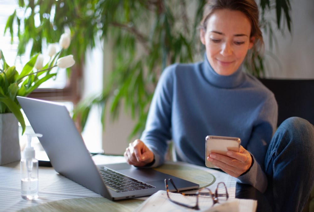 Ung kvinne sitter komfortabelt ved et hjemmekontorbord omgitt av grønne planter og jobber på en laptop mens hun holder en smarttelefon. Hun smiler mens hun ser på telefonen, med briller og en notatbok ved siden av seg på bordet.