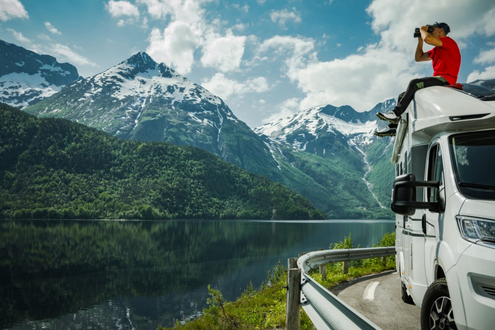 En person i rød t-skjorte står på taket av en hvit bobil og bruker kikkert for å se på landskapet. Bakgrunnen viser et speilblankt fjord omkranset av frodige grønne skoger og snødekte fjelltopper.