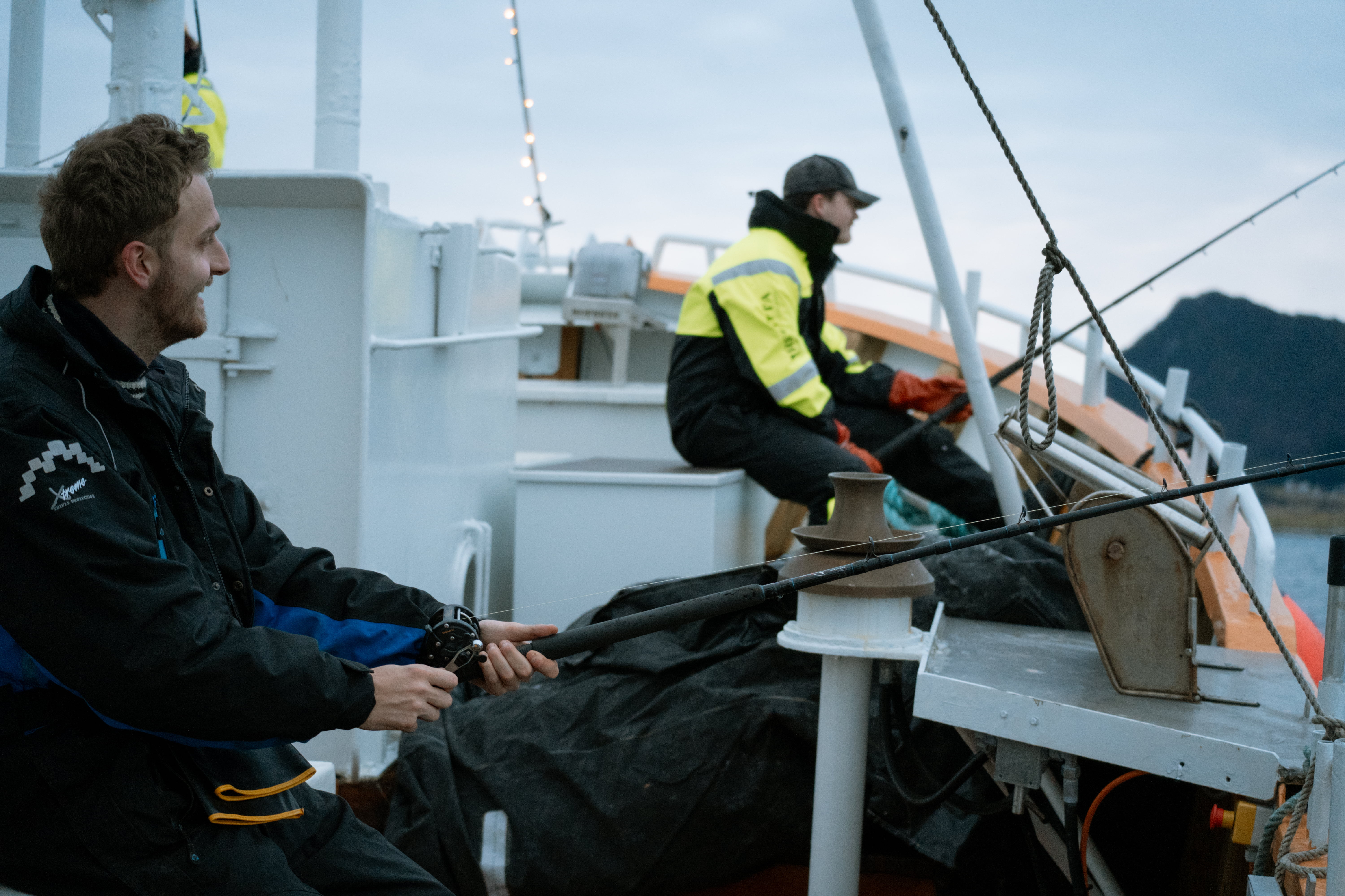 To personer fisker fra en båt med fjell i bakgrunnen.