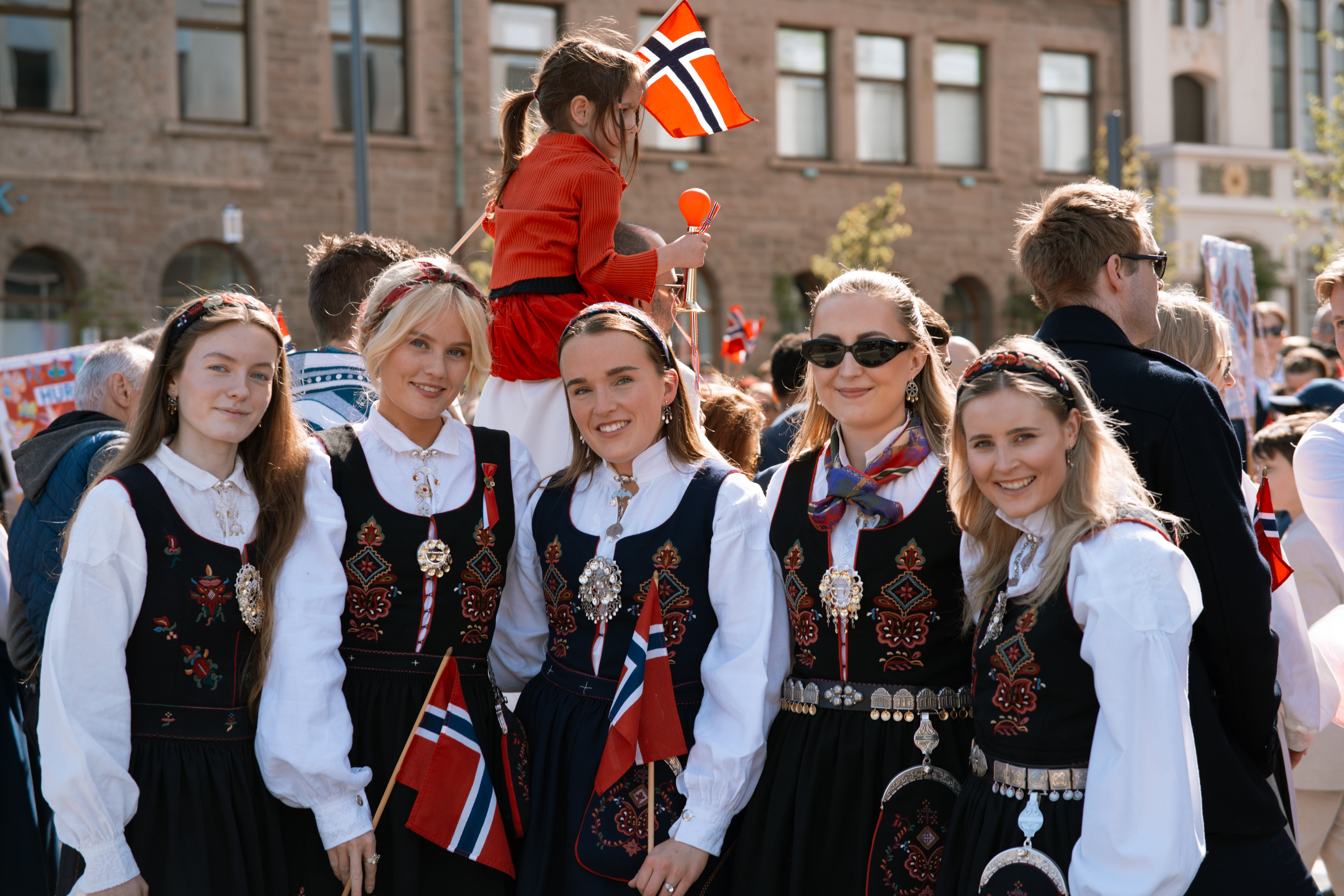 Fem jenter som står ved siden av hverandre foran de som ser på 17. mai-toget.